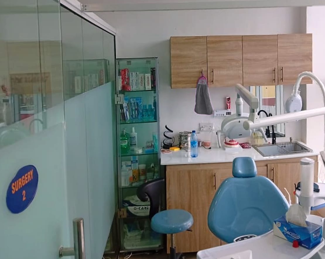 Surgery 2 at Vinix Dental: a blue dental chair and stool beside wooden cupboards, a sink and a glass case of toothbrushes and toothpaste